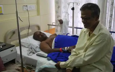A friend sits beside a patient recovering in a hospital bed in India